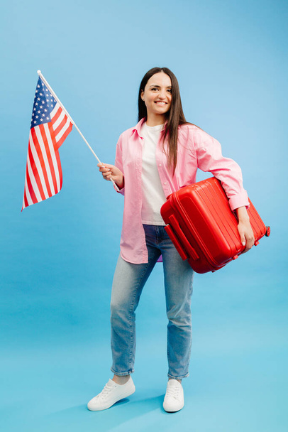 Γυναίκα τουρίστρια με ταξιδιωτική βαλίτσα και σημαία ΗΠΑ, στέκεται απομονωμένη σε μπλε φόντο στούντιο. - Φωτογραφία, εικόνα