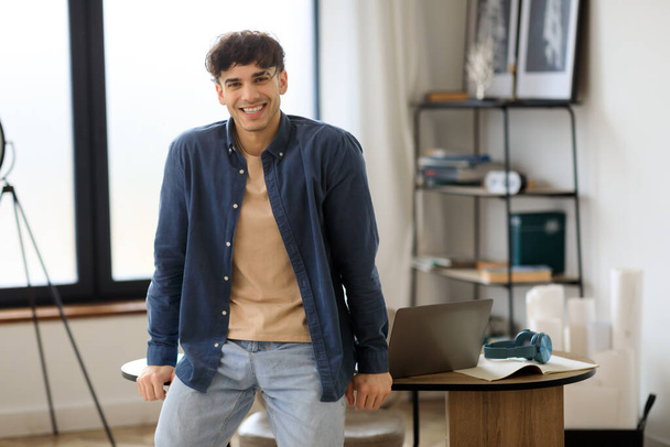 Cheerful Young Businessman Posing Smiling To Camera Sitting On Desk Near Laptop, Smiling To Camera. Shot Of Positive Business Professional Guy In His Modern Office. Career Success Concept - Foto, Imagem