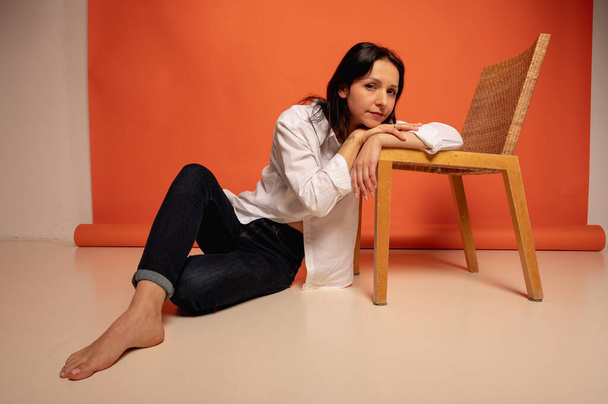 Foto de larga duración de la atractiva mujer de negocios elegante sentada en el suelo apoyada en la silla, el estado de ánimo coqueta, el espacio en blanco en jeans y camisa blanca sobre fondo naranja, lugar para el texto - Foto, imagen