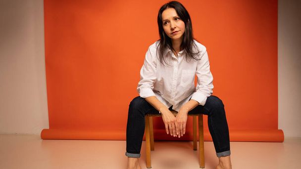 business portrait of a young beautiful girl about 40 years old, cute, emotionally pensive sitting on a wooden chair, on a bright orange background, gesturing with her hands, shot with copy space - Фото, зображення