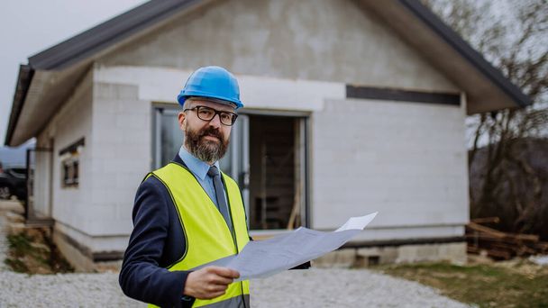 Mature businessman controlling blueprints from an unfinished house. - Fotó, kép