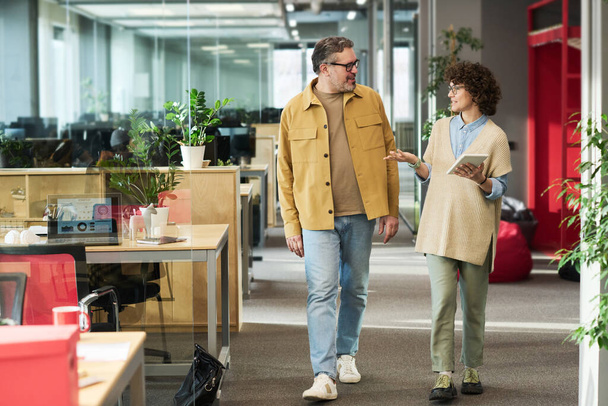 Jovem empresária com tablet olhando para colega masculino maduro durante a conversa, enquanto ambos caminhando ao longo do escritório openspace - Foto, Imagem