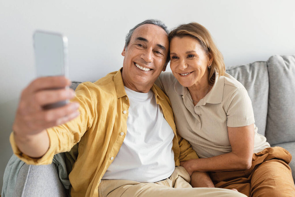 Χαμογελώντας ηλικιωμένος άνδρας και κυρία βγάζοντας selfie στο smartphone, αναπαύεται στον καναπέ στο σπίτι και χαμογελώντας στην κάμερα. Σχέση και αγάπη, φωτογραφία για κοινωνικό δίκτυο και blog - Φωτογραφία, εικόνα