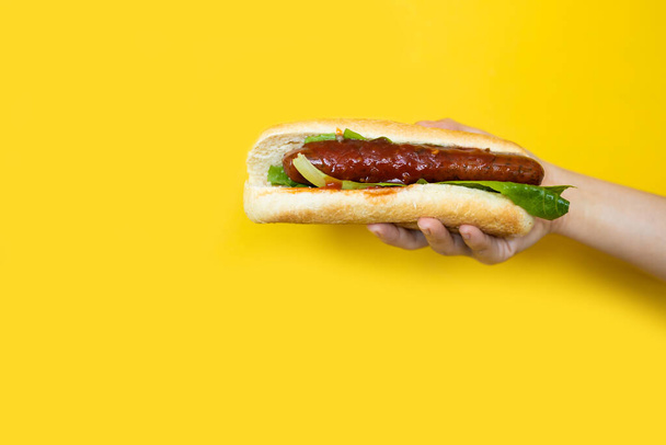 La main tient un hot dog avec saucisse, laitue, ketchup sur fond jaune - Photo, image
