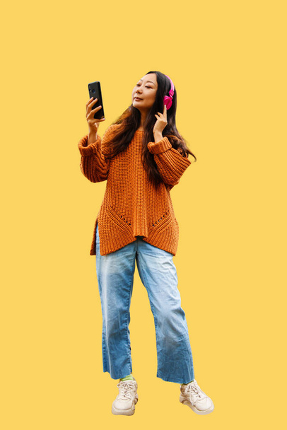 Focalizzato giovane asiatica femmina in maglione indossare ascoltare musica in cuffia e la navigazione cellulare su sfondo giallo - Foto, immagini