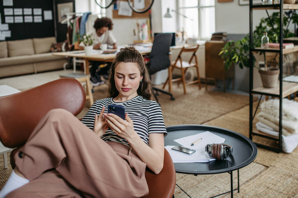 Young woman having break at work, scrolling her smartphone. - Фото, изображение