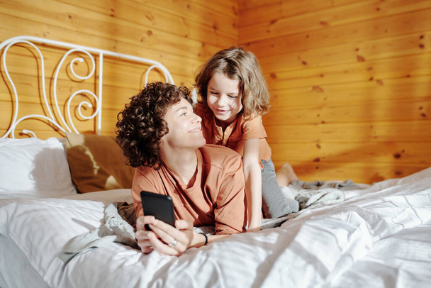 Szczęśliwa młoda kobieta ze smartfonem patrząc na jej słodkiego synka, jednocześnie relaksując się na łóżku i przewijając posty online - Zdjęcie, obraz