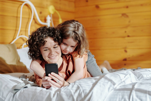 Leuke jongen omarmen zijn moeder met mobiele telefoon, terwijl zowel ontspannen op bed in het weekend in de ochtend en communiceren in video chat - Foto, afbeelding