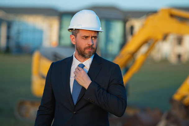 Construction business owner. Man in suit and hardhat halmet at building construction site. Civil engineer worker in front of house near excavator. Architect, supervisor, investor, construction manager - Foto, Bild