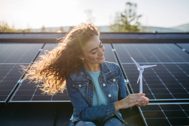 Portrait of young woman, holding model of a wind turbine on roof with solar panels. - Valokuva, kuva