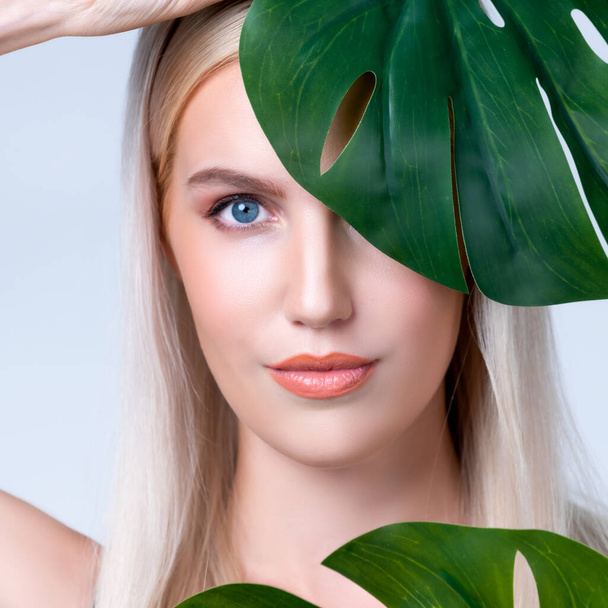 Closeup facial portrait personable woman with perfect smooth makeup holding green monstera leaves and cover her face as natural healthy skincare treatment. Tropical nature and beauty concept - Fotó, kép