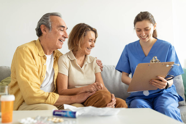 Wizyta u pacjenta. Pielęgniarka rozmawiająca z parą seniorów, trzymająca notatnik w rozmowie ze starszym mężczyzną i kobietą, przynosząca wyniki badań lekarskich - Zdjęcie, obraz