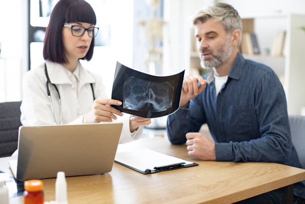 Skilled radiologist in white uniform and eyeglasses explaining visible changes on x-ray image. Anxious man dressed in jeans and denim shirt carefully listening to physicians instructions. - Foto, afbeelding