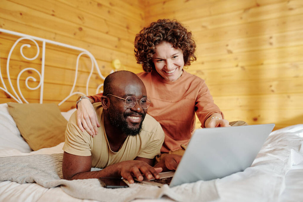 Kocasına sarılmış gülümseyen ve dizüstü bilgisayarı işaret eden genç bir kadın hem yatakta dinleniyor hem de online video izliyor. - Fotoğraf, Görsel