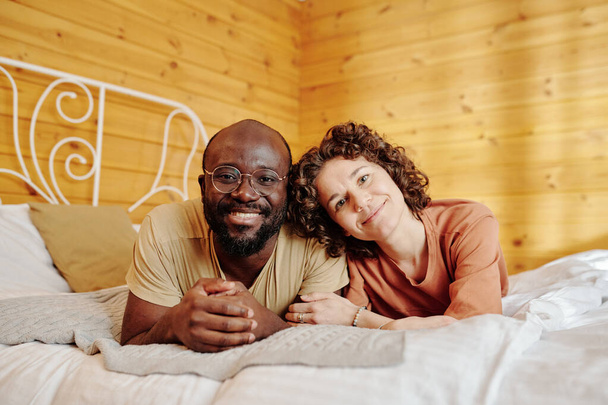 Happy young intercultural couple in pajamas relaxing on comfortable double bed after sleep in the morning and looking at camera - Фото, зображення
