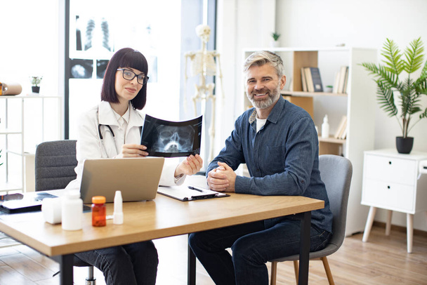 Portrait of happy doctor and patient sitting at table with portable computer in modern medical office and smiling at camera. Qualified radiologist dressed in white lab coat holding x-ray in hands. - 写真・画像