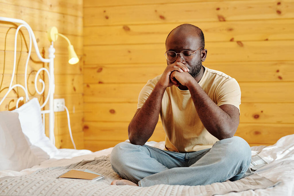 Young tense man in jeans and t-shirt keeping clenched hands by his face while sitting on bed with his legs crossed and thinking - Photo, Image