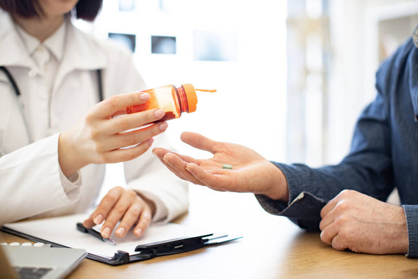 Close up of brunette doctor pouring tablets from orange bottle on patients hand in modern polyclinic. Male person taking pills to reduce symptoms of illness and pain. Concept of health and treatment. - Φωτογραφία, εικόνα