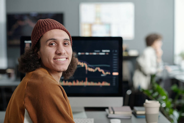 Glücklicher junger IT-Support-Manager blickt in die Kamera, während er am Arbeitsplatz vor dem Computerbildschirm mit Grafikdaten sitzt - Foto, Bild