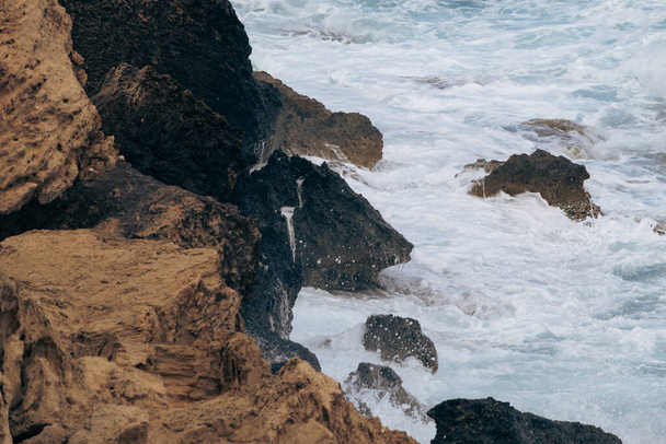 Las olas chocan contra las rocas en la orilla del océano. Agua de mar azul con espuma en la orilla del mar. - Foto, imagen