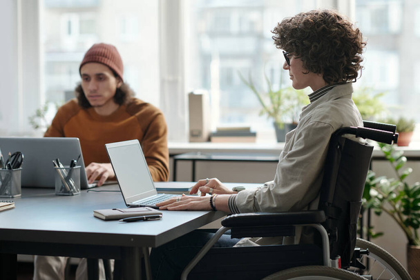 Jonge vrouwelijke econoom met een handicap zit op een rolstoel voor laptop en analyseert online financiële gegevens of het organiseren van werk - Foto, afbeelding