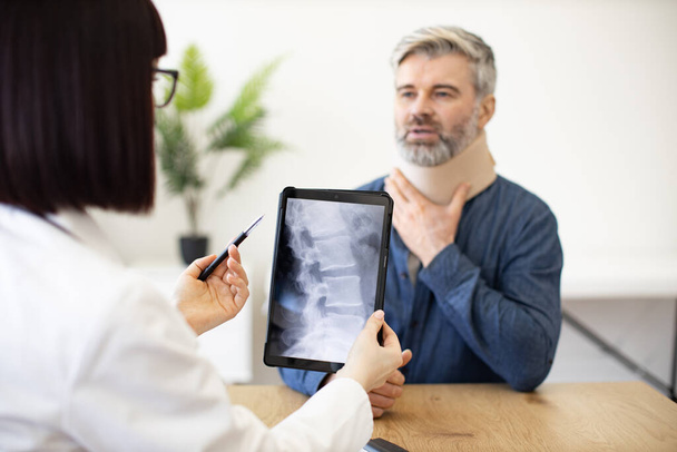Handsome male in neck brace listening to therapist with digital x-ray images while sitting in doctors office. Focused patient with physical sprain visiting family doctor for diagnosis in clinic. - Foto, immagini
