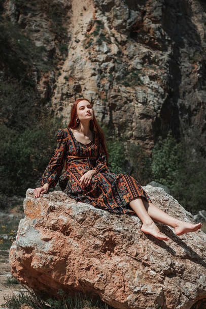 Δυνατή, ελεύθερη, δυνατή, κοκκινομάλλα γυναίκα με ένα έθνικ φόρεμα κοντά σε μια μεγάλη πέτρα. Ένα σύμβολο ακαταλληλότητας. Καριέρα - Φωτογραφία, εικόνα