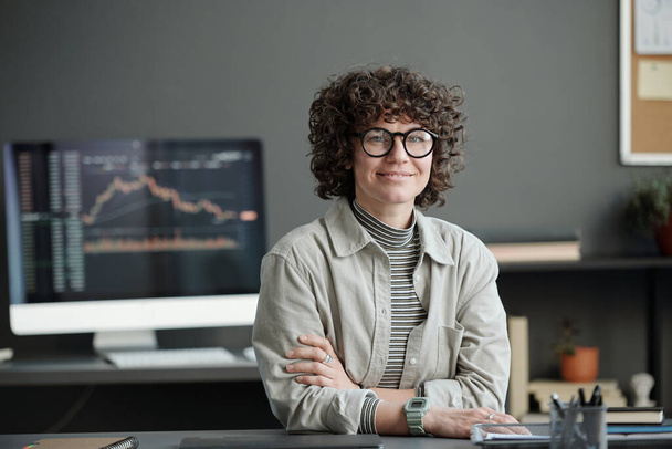 Young curly brunette businesswoman in casualwear and eyeglasses sitting by workplace against computer screen with graph and chart - Photo, image