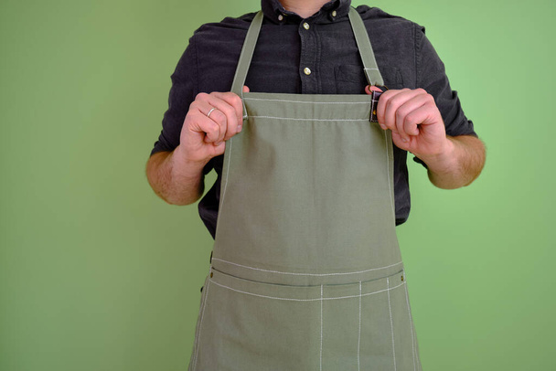 A man in a kitchen apron. Chef work in the cuisine. Cook in uniform, protection apparel. Job in food service. Professional culinary. Green fabric apron, casual stylish clothing. Handsome baker posing - Fotó, kép