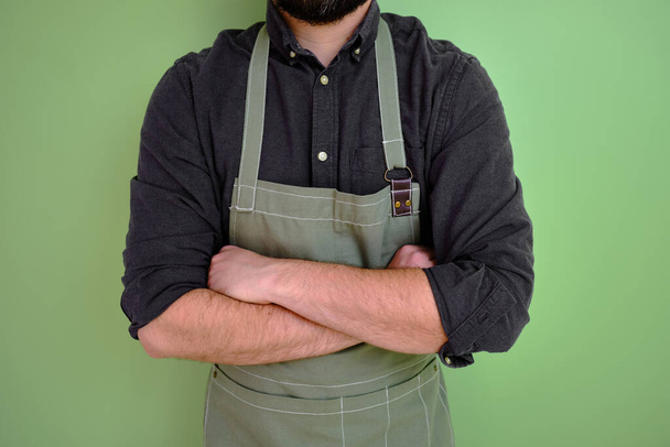 A man in a kitchen apron. Chef work in the cuisine. Cook in uniform, protection apparel. Job in food service. Professional culinary. Green fabric apron, casual stylish clothing. Handsome baker posing - Foto, afbeelding