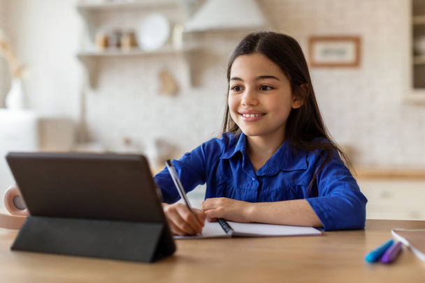 Μαθαίνω. Happy Middle Eastern Schoolgirl κοιτάζοντας ψηφιακή ταμπλέτα και λαμβάνοντας υπόψη, Excelling in Her Studies through Online Education at Home, κάνει Homewotk κάθεται στο γραφείο κοντά στον υπολογιστή - Φωτογραφία, εικόνα