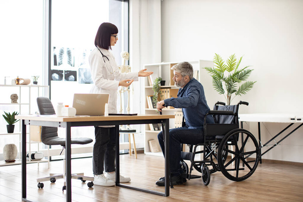 Young woman in lab coat helping mature man with walking cane to stand up from wheelchair in consulting room. Efficient therapist assisting patient in finding balance during recovery from injury. - Photo, image