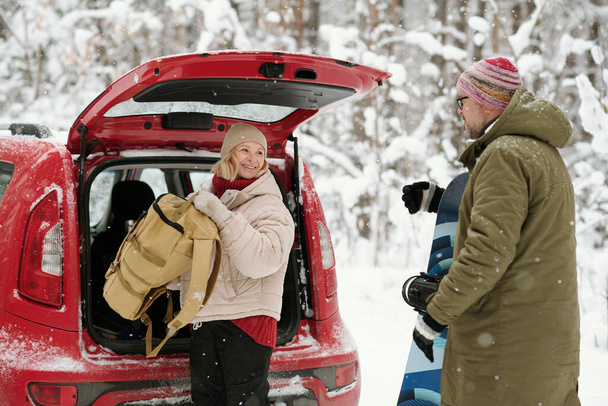 Mujer mayor feliz con mochila mirando a su marido con snowboard mientras saca el equipaje del maletero del coche - Foto, imagen