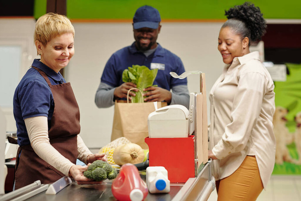 Glückliche reife Verkäuferin steht im Supermarkt an der Kasse und bedient junge afroamerikanische Konsumentinnen - Foto, Bild