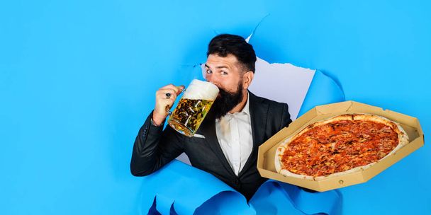 Brodaty facet ze smaczną pizzą pijący kubek piwa. Czas na pizzę. Zadowolony człowiek z pyszną pizzą i zimnym piwem patrzący przez papierową dziurę. Szybkie żarcie. Włoskie jedzenie. Dostawa pizzy. Biznesowy obiad - Zdjęcie, obraz