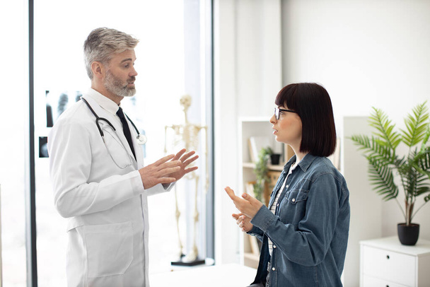 Attractive young woman holding hand on throat while describing health condition to bearded man in lab coat. Confident therapist giving consultation about illness to female patient at modern hospital. - Foto, Imagem