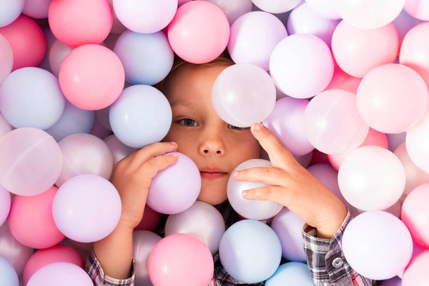 Little boy in a pool filled with plastic balls. Child peeking from pastel multi colored plastic balls for kids. top view. - Photo, Image