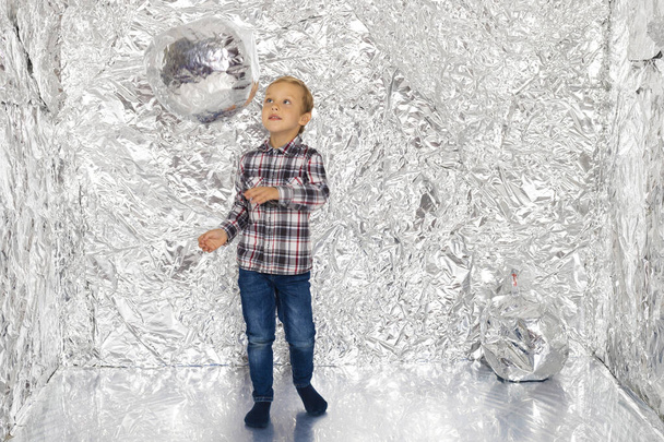 Мальчик играет в комнате из фольги. Ребенок в отражающей блестящей комнате играет с мячом. - Фото, изображение