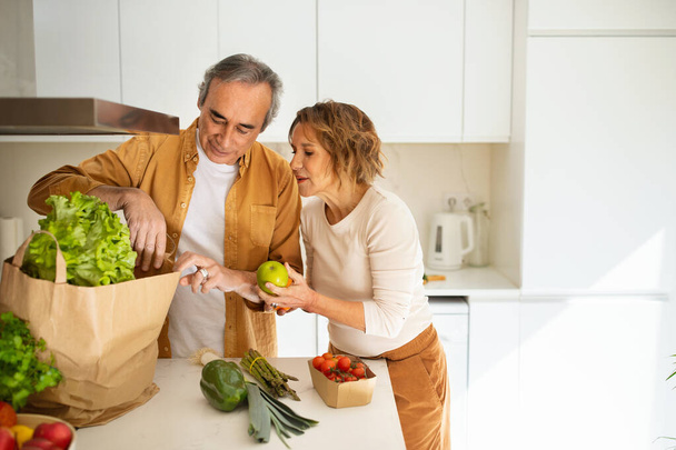 Elderly spouses unpacking paper bag with fresh vegetables and fruits in kitchen, ordering food or coming home from supermarket, free space. Retirement and healthy eating - Фото, изображение