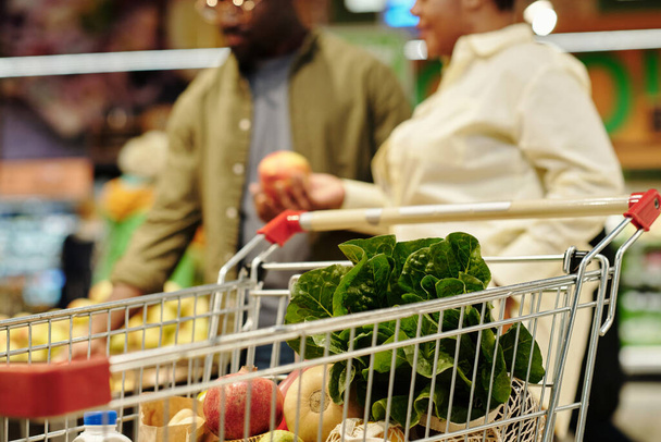 Ostoskori tuoreita hedelmiä ja vihanneksia vastaan nuori pari valitsemalla omenat seistessään ruokakaupassa - Valokuva, kuva