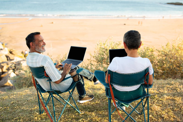 Sonho Freelance. Visão traseira do casal sênior usando laptops Computadores sentados em cadeiras na praia perto do oceano ao ar livre. Cônjuges maduros navegando na Internet de férias, desfrutando de trabalho on-line de qualquer lugar - Foto, Imagem