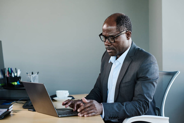 Grave maturo uomo d'affari afroamericano in abbigliamento formale e occhiali da vista utilizzando il computer portatile mentre seduto alla scrivania nel suo ufficio - Foto, immagini
