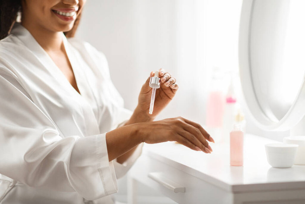 Smiling african american woman applying serum on her hand at home, attractive black female making daily beauty routine while sitting at dressing table in bedroom, cropped shot with free space - Foto, Imagem