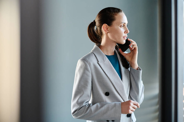 Junge seriöse Geschäftsfrau in Formalbekleidung hält Smartphone am Ohr, während sie im Büro vor dem Fenster steht und Untergebene anruft - Foto, Bild