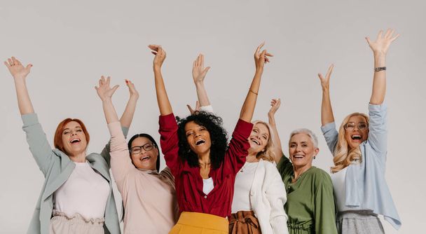 Multi-ethnic group of mature women keeping arms raised and smiling against grey background - 写真・画像
