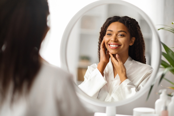 Concepto de cuidado de la piel. Hermosa mujer negra joven mirando en el espejo y tocando su cara, atractiva mujer afroamericana disfrutando de una piel suave y suave después de tratamientos de belleza, enfoque selectivo - Foto, Imagen