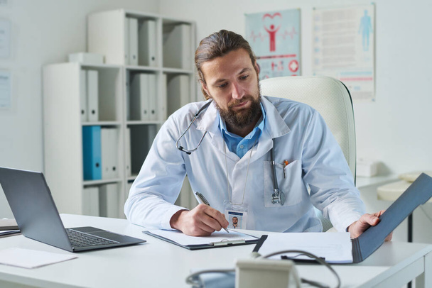 Молодой виртуальный медицинский ассистент просматривает документы и заполняет медицинскую форму пациента, сидя на рабочем месте в клиниках - Фото, изображение