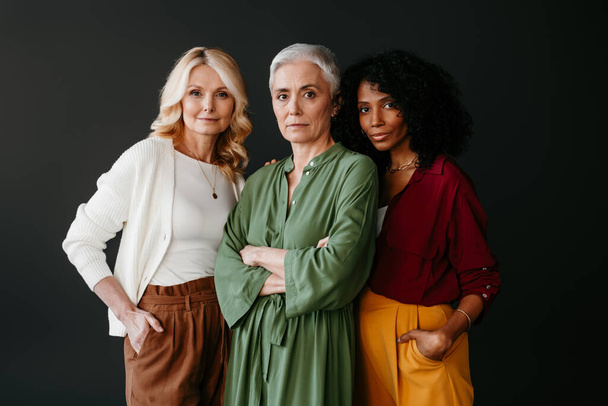 Three confident mature women looking at camera while bonding against grey background - Фото, зображення