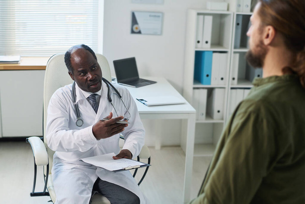 Confident mature male doctor with medical document talking to young patient sitting in front of him during consultation in clinics - Photo, Image
