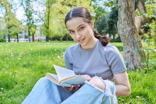 Teenage girl středoškolačka čtení knihy sedí na trávě v parku. Usmívající se mladá žena dívající se do kamery - Fotografie, Obrázek